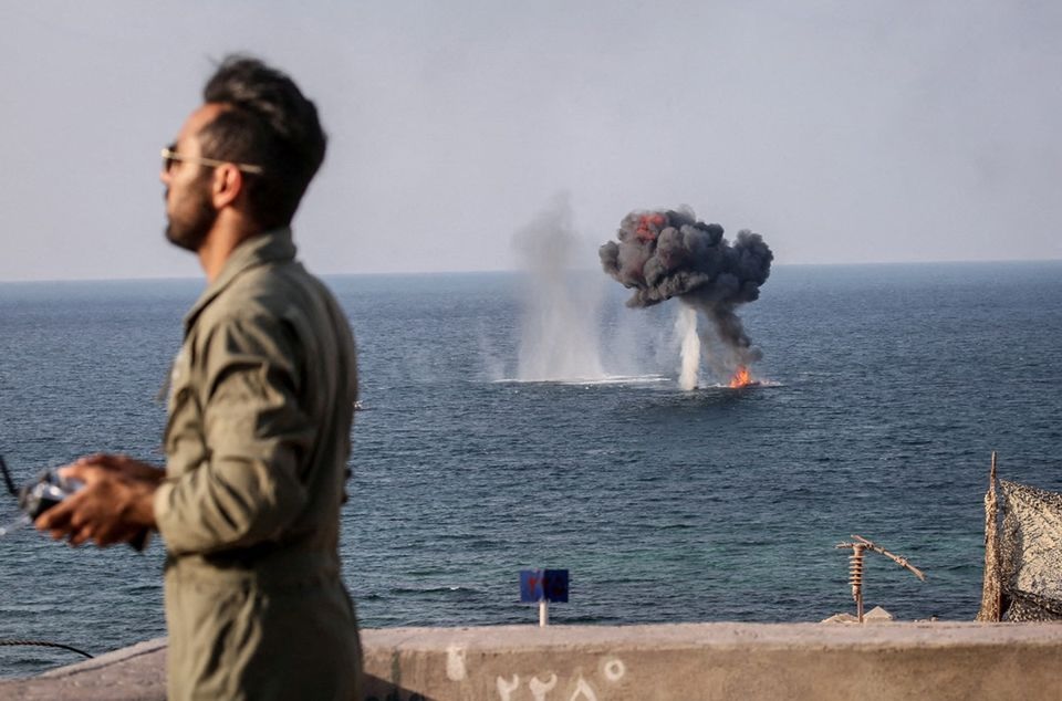 Sebuah ledakan terlihat di permukaan air di belakang seorang anggota Korps Pengawal Revolusi Islam (IRGC) selama latihan militer bersama yang disebut 'Nabi Besar 17' di barat daya Iran, dalam gambar ini diperoleh pada 22 Desember 2021. Foto: Reuters.