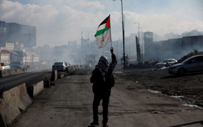 Seorang pria berjalan dengan bendera Palestina, ketika warga Palestina bentrok dengan pasukan Israel selama protes terhadap apa yang dikatakan penyelenggara sebagai tindakan Israel untuk menahan mayat tahanan, di dekat Qalandia di Tepi Barat yang diduduki Israel 27 Desember 2022. Foto: Reuters/Mohamad Torokman.