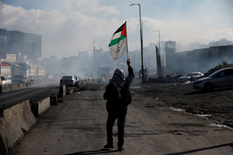 Seorang pria berjalan dengan bendera Palestina, ketika warga Palestina bentrok dengan pasukan Israel selama protes terhadap apa yang dikatakan penyelenggara sebagai tindakan Israel untuk menahan mayat tahanan, di dekat Qalandia di Tepi Barat yang diduduki Israel 27 Desember 2022. Foto: Reuters/Mohamad Torokman.